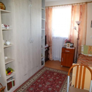widok pokoju jednoosobowego (można dostrzec łóżko, krzesło, telewizor, okno, szafy, małą lodówkę, szafeczkę przyłóżkową, stół))
