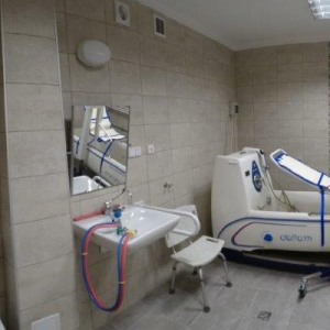 widok łazienki z wanną do hydromasażu z dźwigiem wspomagającym oraz urządzeniami sanitarnymi (zlew z lustrem regulowanym, sedes toaletowy) i poręczami wspomagającymi.