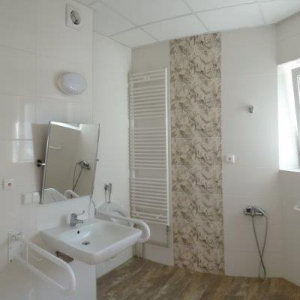 widok łazienki z urządzeniami sanitarnymi (prysznic, zlew z lustrem regulowanym, pisuar) i poręczami wspomagającymi